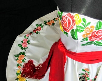 May include: White dress with colorful floral embroidery, featuring red, yellow, and orange roses with green leaves. A red sash with fringe detail accents the waist. The dress is likely for a special occasion.