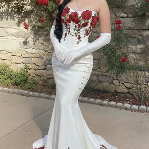 May include: A strapless white gown with red rose embroidery. The dress features a mermaid silhouette, long white gloves, and decorative gold accents. The roses are also embroidered on the train of the dress. The background is a stone wall with red roses.