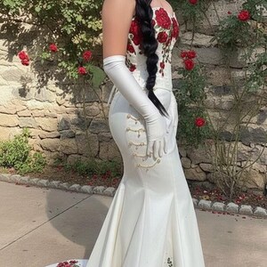 May include: A white satin gown with red rose embroidery. The dress features a fitted bodice, a mermaid silhouette, and a long train. Long white gloves and a braided hairstyle complete the look.