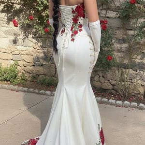 May include: A white strapless wedding gown with a mermaid silhouette. The bodice and train are adorned with red rose embroidery. The back features a corset closure. Long white gloves complete the look.