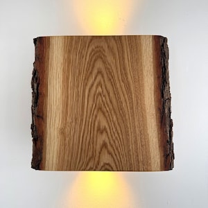 Puede incluir: Aplique de pared rectangular de madera con borde natural. La veta de la madera es visible y los bordes tienen una textura rugosa, similar a la corteza. El aplique emite una luz cálida y amarilla arriba y abajo.
