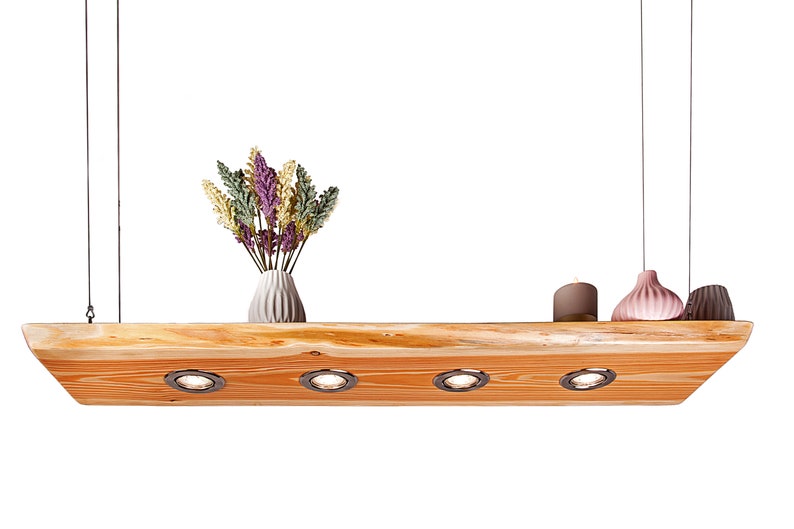 May include: A wooden ceiling light fixture with four integrated spotlights. The fixture is suspended from the ceiling by two cords. The fixture is made of natural wood with a rustic finish. The fixture is decorated with a vase of dried flowers and two decorative ceramic objects.
