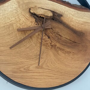 May include: A round wooden clock with a natural edge and a dark metal frame. The clock face is made from a light-colored wood with visible grain and knots. The clock hands are a darker brown color.