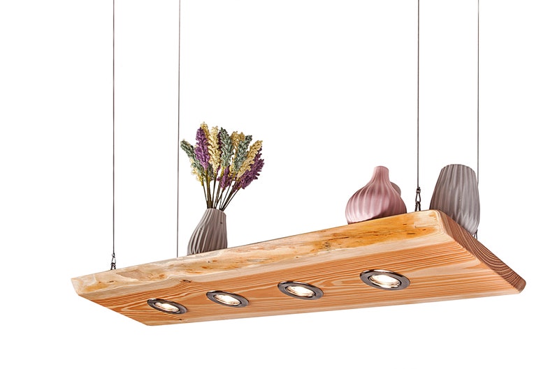 May include: A wooden ceiling light fixture with four integrated spotlights. The fixture is suspended from the ceiling by four wires. The wood is a natural, light brown color. There are three decorative vases on the fixture, one pink, one gray, and one beige. The fixture is lit and the light is shining on the vases.