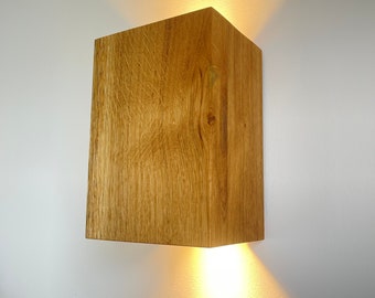 Puede incluir: Una lámpara de pared cuadrada de madera con una luz blanca cálida que brilla desde la parte inferior. La lámpara está hecha de madera natural con un acabado marrón claro.