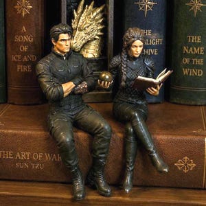 May include: Two detailed figurines of a man and a woman sitting on a brown book titled The Art of War. The man wears a black jacket and pants, holding a gold sphere. The woman wears black armor and reads a book. Behind them is a gothic arch with a dragon.