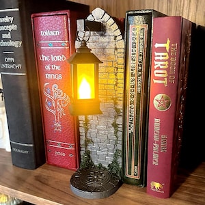 May include: A book nook insert featuring a gray stone wall with a hanging black lantern that emits a warm yellow light. The structure includes a circular base with a floral pattern and green moss accents, positioned between several hardcover books.