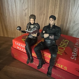 May include: Two detailed character figurines sitting on red books. One figure has braided hair, wears dark armor, and holds a small sphere and an open book. The other figure has dark hair, wears a black long-sleeved shirt with arm tattoos, and sits with arms crossed.
