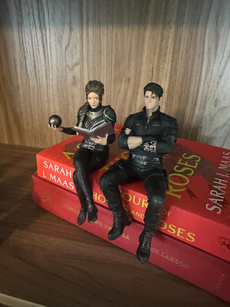 May include: Two detailed character figurines sitting on red books. One figure has braided hair, wears dark armor, and holds a small sphere and an open book. The other figure has dark hair, wears a black long-sleeved shirt with arm tattoos, and sits with arms crossed.