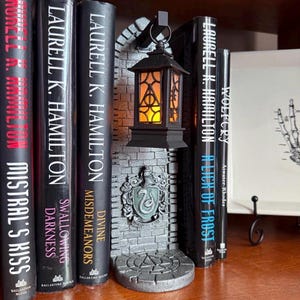 May include: A gray stone-textured bookend featuring a hanging black lantern with a glowing orange light and a crest displaying a snake. The lantern panels show the Deathly Hallows symbol. The bookend is positioned between several black books.