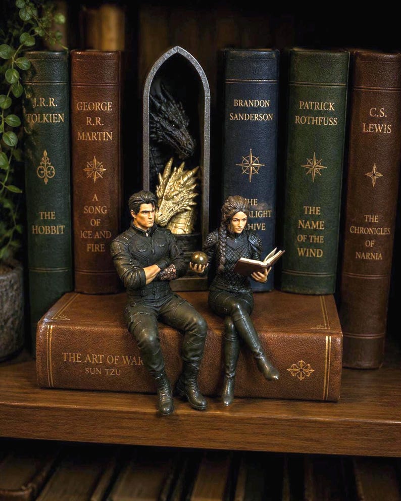 May include: Two detailed figurines of a man and a woman sitting on a brown book titled The Art of War. The man wears a black jacket and pants, holding a gold sphere. The woman wears black armor and reads a book. Behind them is a gothic arch with a dragon.