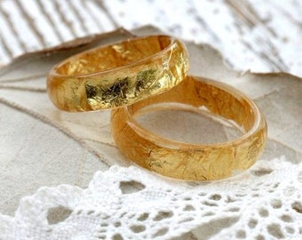 Könnte beinhalten: Zwei goldene Ringe mit einer strukturierten Oberfläche und goldenen Blatt-Akzenten. Die Ringe liegen auf einem weißen Spitzenhintergrund.