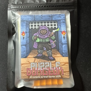 Pode incluir: Uma bolsa preta e selável contendo um jogo. A arte da capa do jogo apresenta gráficos pixelizados de uma figura vestida com uma túnica em um cenário de masmorra, com o texto "PUZZLE DUNGEON" e "All contents by Brian Garber 2025".