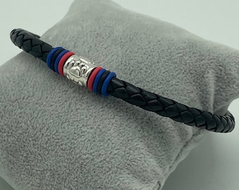 Può includere: Bracciale in pelle intrecciata nera con dettagli argentati. Il bracciale presenta fasce rosse e blu e una chiusura decorativa argentata. Il bracciale è esposto su una superficie grigia e morbida.