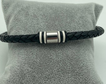 Può includere: Un braccialetto in pelle intrecciata nera con chiusura magnetica argentata e dettagli bianchi. Il braccialetto è presentato su un cuscino di peluche grigio, evidenziandone il design. Un accessorio per l'uso quotidiano.