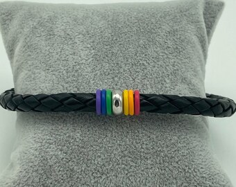 Può includere: Bracciale in pelle intrecciata nera con un accento di perline color arcobaleno. Il bracciale presenta una serie di perline colorate nei colori dell'arcobaleno, tra cui rosso, arancione, giallo, verde, blu e viola. Il bracciale è esposto su un cuscino grigio.