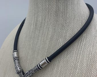 Può includere: Una collana in gomma nera con dettagli argentati a forma di testa di drago e un ciondolo rettangolare nero. La collana è esposta su un manichino.