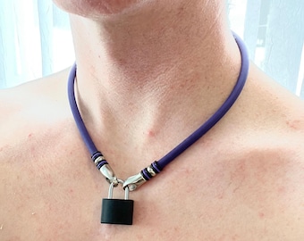 Può includere: Una collana in corda di gomma viola con un ciondolo a forma di lucchetto nero opaco. La corda è fissata sul davanti con elementi in metallo argentato, inclusi due piccoli anelli argentati su ciascun lato collegati all'arco del lucchetto.