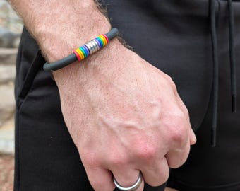 Può includere: Un braccialetto in silicone nero opaco con una fila centrale di piccole perline a righe color arcobaleno alternate a distanziatori in argento metallizzato. Un anello in metallo liscio color argento indossato sull'anulare.