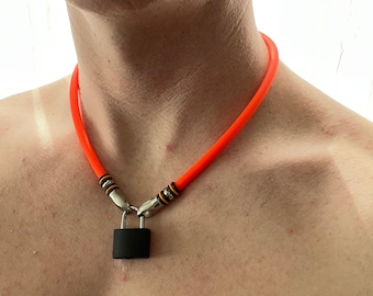 Può includere: Una collana a tubo in silicone color arancione neon brillante con un ciondolo a forma di lucchetto nero opaco. La collana è fissata con terminali in metallo argentato con due sottili anelli neri, che collegano il tubo al lucchetto.