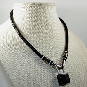 Può includere: Una collana in pelle nera con un ciondolo a forma di lucchetto argentato. La collana è decorata con cuciture bianche e accenti argentati.