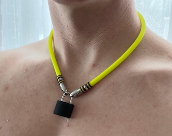 Può includere: Una collana in tubo di gomma giallo neon con un ciondolo a forma di lucchetto nero opaco. La collana è fissata con connettori in metallo argentato che presentano anelli neri e gialli dove il tubo incontra l'arco del lucchetto.