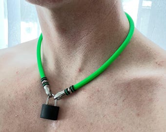 Può includere: Una collana girocollo in gomma verde neon brillante con connettori in metallo argentato e un ciondolo a forma di lucchetto nero opaco appeso al centro. La fascia liscia e flessibile aderisce al collo con un meccanismo di chiusura in metallo.