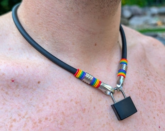 Può includere: Una collana girocollo in gomma nera con un piccolo lucchetto nero come ciondolo. La collana presenta dettagli in metallo color argento e segmenti di perline color arcobaleno posizionati su entrambi i lati del punto di attacco del lucchetto.