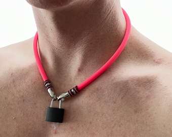 Può includere: Una collana girocollo in gomma rosa neon con un lucchetto nero opaco come ciondolo. La collana è fissata con terminali in metallo argentato che si collegano al lucchetto, con piccoli anelli in gomma nera che accentuano i componenti metallici.
