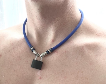 Può includere: Una collana girocollo in gomma blu con un ciondolo a forma di lucchetto nero opaco. La collana presenta terminali in metallo argentato con dettagli ad anello neri e blu che si collegano al lucchetto, creando un accessorio audace.
