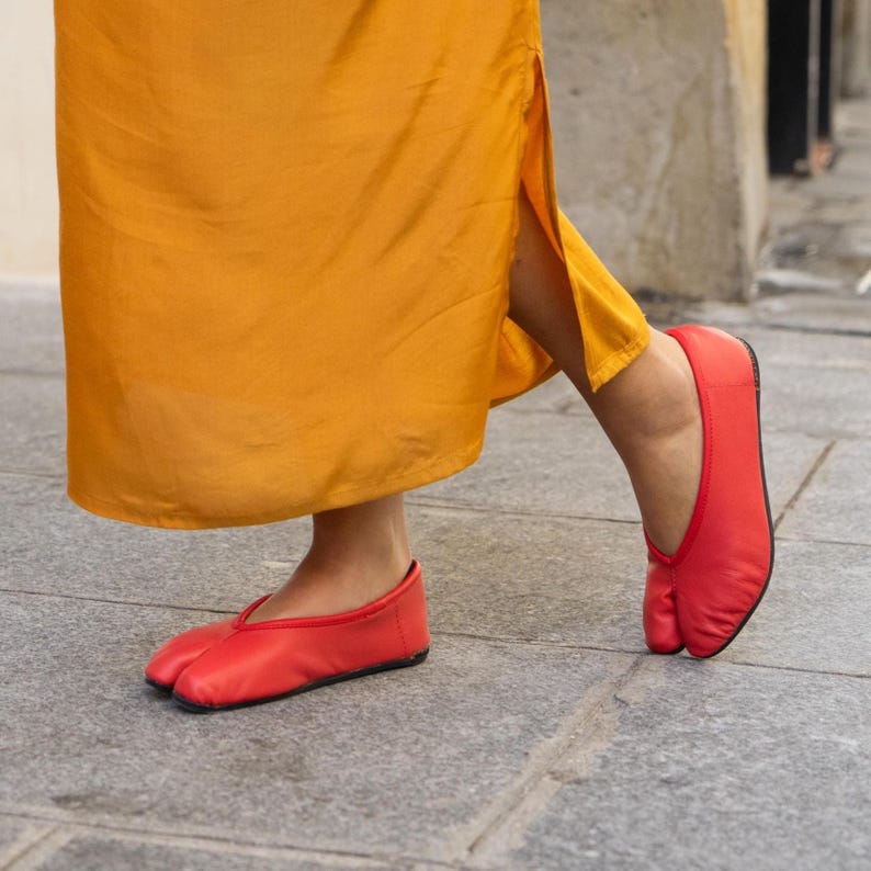 May include: A pair of red split-toe ballet flats. The shoes are made of a smooth material and feature a low profile design. The flats are worn with a long, flowing, yellow garment.