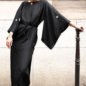 May include: A black dress with wide sleeves and white button details. The dress has a tie waist and is paired with beige boots and a brown hat. The dress is a midi length and appears to be made of a smooth fabric.