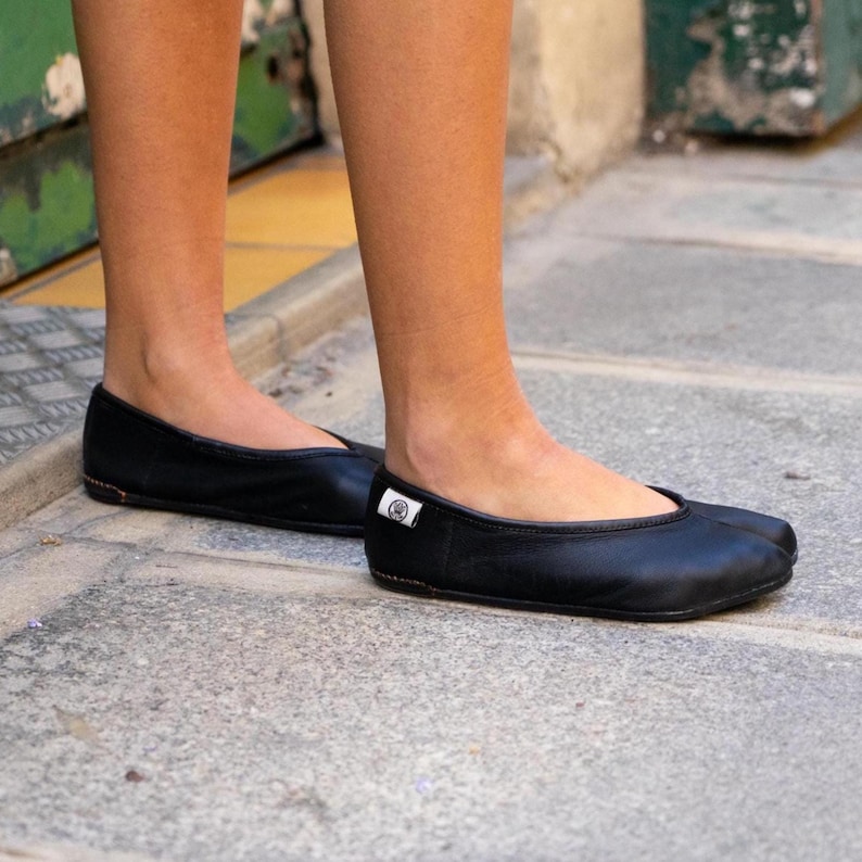 May include: A pair of black leather ballet flats. The shoes have a rounded toe and a small, flat heel. A small white tag with a logo is visible on the side of one shoe. The shoes are worn by a person.