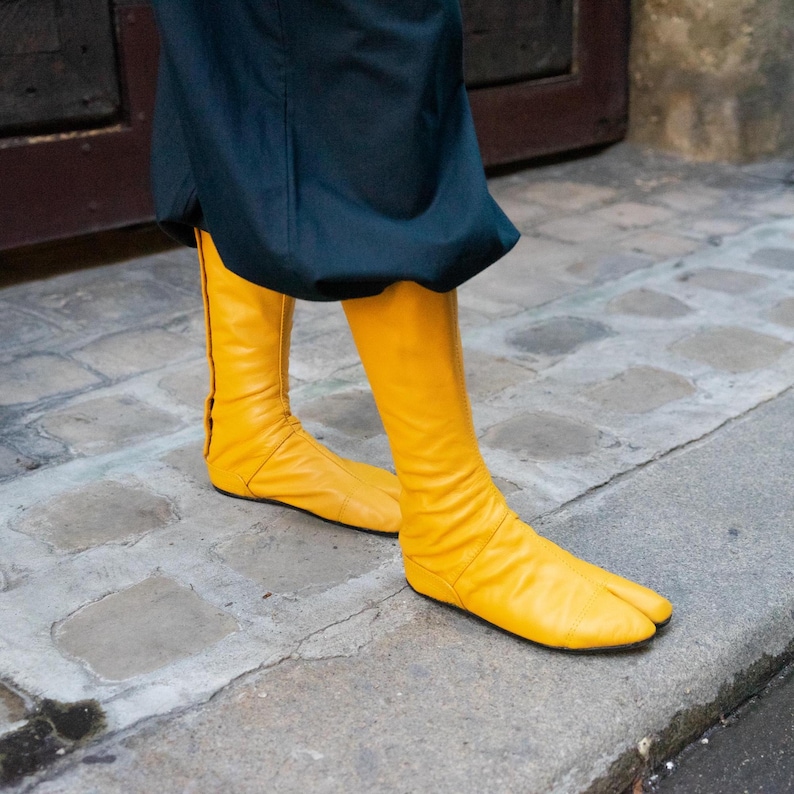 May include: A pair of yellow leather boots with a split toe design. The boots are knee-high and have a zipper on the back.