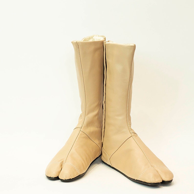May include: A pair of tan leather tabi boots with split toes. The boots are knee-high and have a simple design.