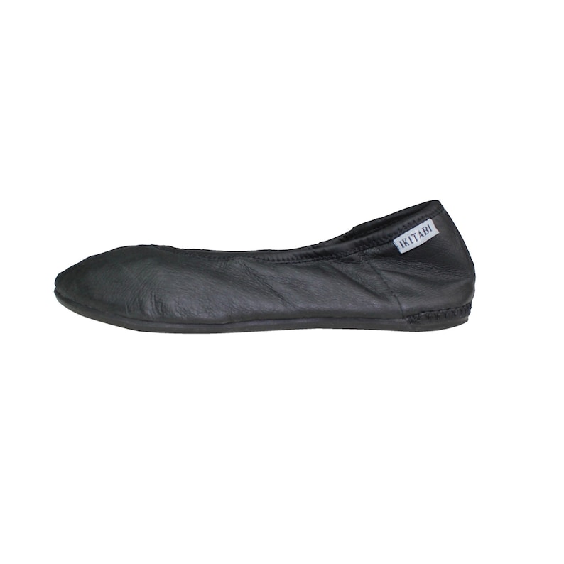 May include: Black leather ballet flat with a rounded toe. The shoe has a stitched sole and a small white tag that reads "IKITABI". The shoe is designed for comfort and flexibility.