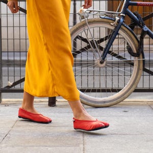 May include: A person wearing a mustard yellow dress and red ballet flats walks on a sidewalk next to a blue bicycle. The dress has wide sleeves. The flats are a bright red color.