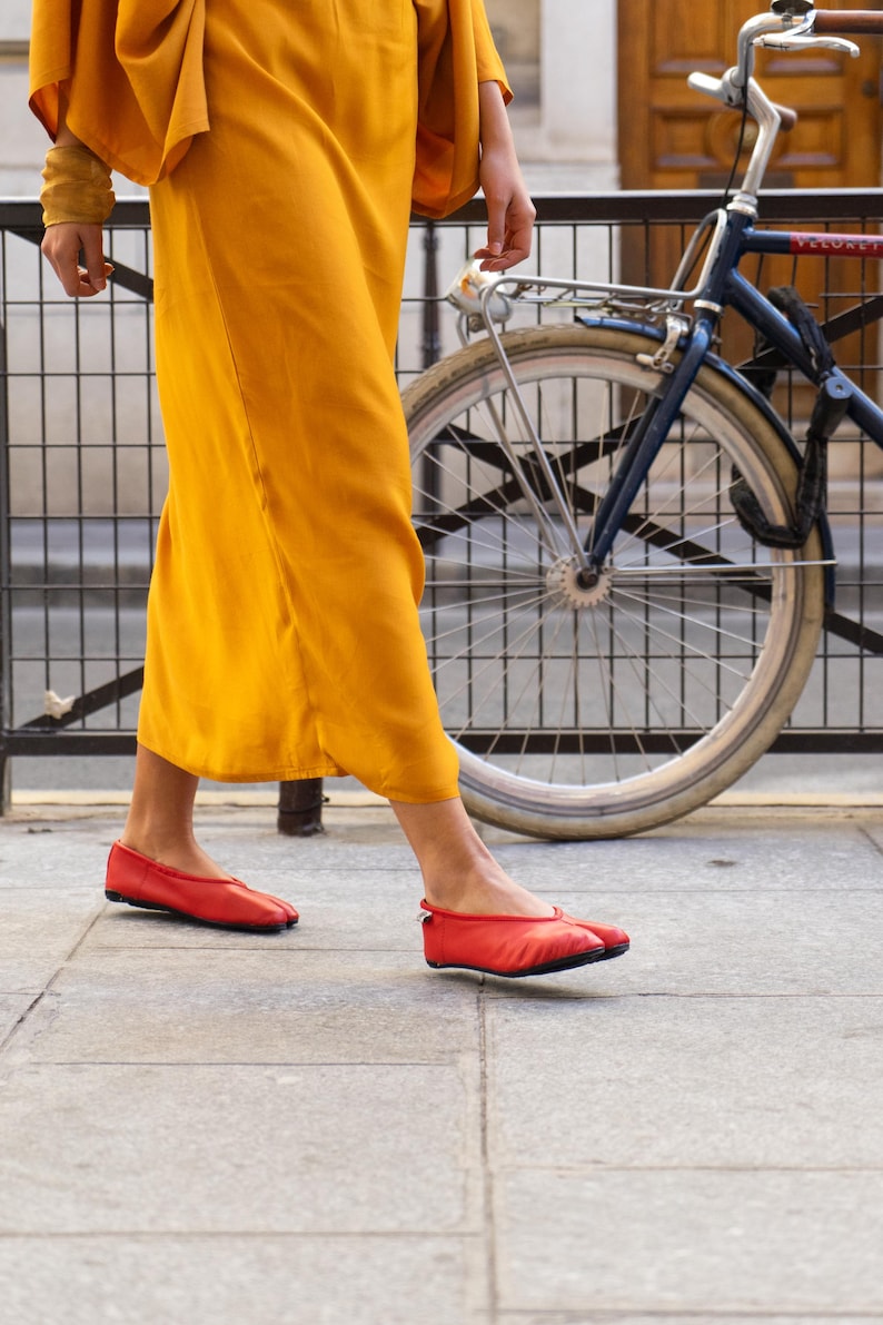 May include: A person wearing a mustard yellow dress and red ballet flats walks on a sidewalk next to a blue bicycle. The dress has wide sleeves. The flats are a bright red color.