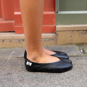 May include: Black leather ballet flats with a rounded toe. The flats have a small, white tag with a circular logo on the side. The shoes are worn on a stone surface, with a red and green background.
