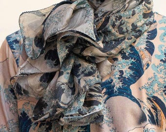 May include: A lightweight scarf and a short-sleeved shirt featuring a repeating print of the Great Wave off Kanagawa. The design displays dark blue crashing waves with white foam against a muted beige background, rendered in a fluid fabric.