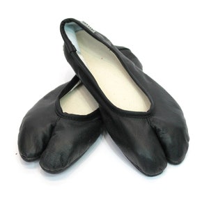 May include: Pair of black split-toe shoes, likely for martial arts or dance. The shoes have a soft, leather-like appearance with a split between the big toe and the other toes. The interior is white, and the shoes are set against a white background.
