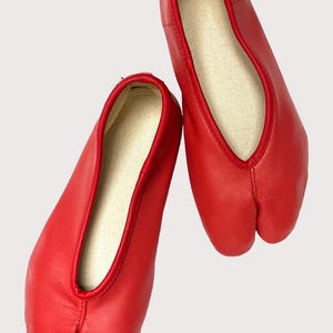 May include: A pair of bright red split-toe ballet flats. The shoes have a smooth leather exterior and a contrasting beige interior. The design features a split between the big toe and the other toes, and a low-cut vamp.