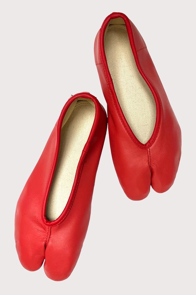 May include: A pair of bright red split-toe ballet flats. The shoes have a smooth leather exterior and a contrasting beige interior. The design features a split between the big toe and the other toes, and a low-cut vamp.