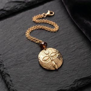 May include: Gold-toned pendant necklace featuring a detailed mask design. The round pendant has etched facial features and floral patterns. A brown bead accents the chain, which is attached to a clasp.