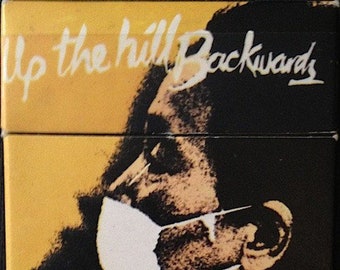 May include: A black and white album cover with a man wearing a white face mask. The text "Up the Hill Backwards" is in white on a yellow background. The text "DAVID BOWIE" is in yellow at the bottom of the image.