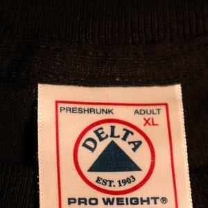 May include: White tag with black text that reads "Preshrunk Adult XL" and a red circle with a blue triangle inside that reads "Delta Est. 1903" and "Pro Weighto". The tag also reads "Assembled in Mexico of U.S. Components Care & Content on Reverse".