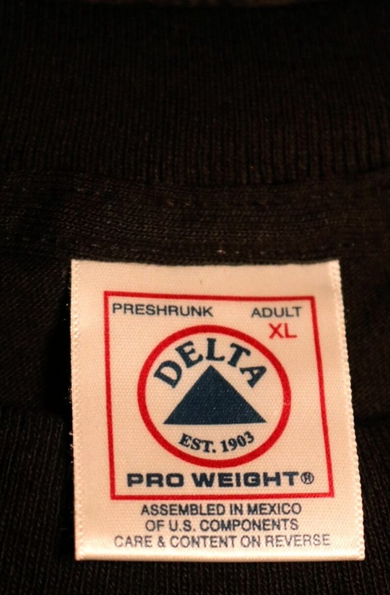 May include: White tag with black text that reads "Preshrunk Adult XL" and a red circle with a blue triangle inside that reads "Delta Est. 1903" and "Pro Weighto". The tag also reads "Assembled in Mexico of U.S. Components Care & Content on Reverse".
