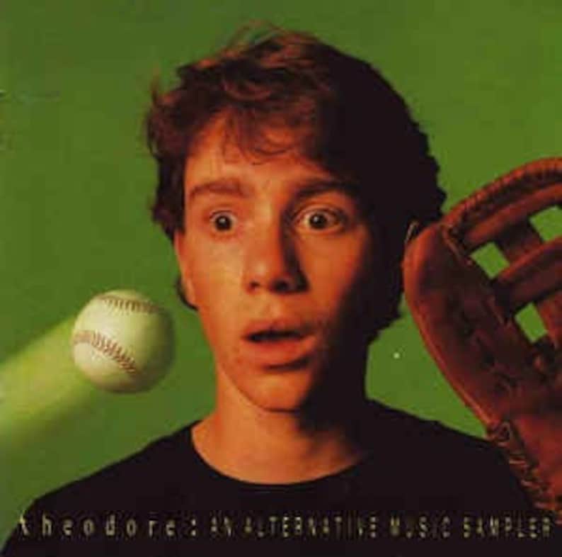 May include: A young person with a surprised expression is wearing a black shirt and looking at a baseball flying towards them. A baseball glove is in the background.
