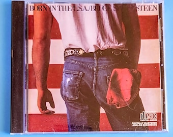Puede incluir: Caja de CD del álbum "Born in the U.S.A." de Bruce Springsteen. La portada muestra a un hombre en vaqueros y camiseta blanca, con una bandera americana de fondo. El CD está en una caja de plástico transparente.