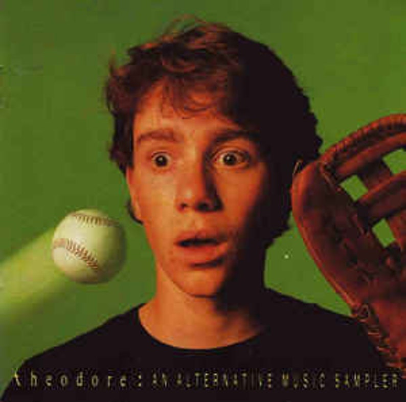 May include: A young person with a surprised expression is looking at a baseball flying towards them. The person is wearing a black shirt and a brown baseball glove is in the background. The text "Theodore: An Alternative Music Sampler" is at the bottom of the image.
